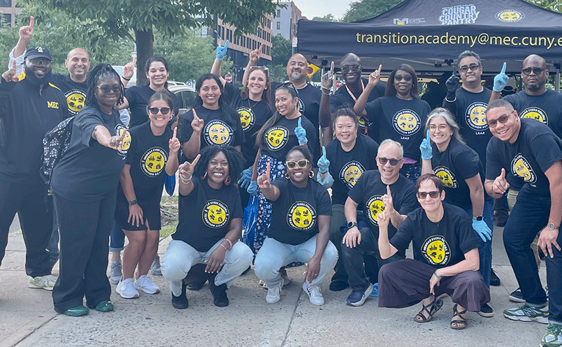 Group of volunteers from Baruch College's Online Executive Doctor of Education in Higher Education Administration program wearing matching MEC T-shirts pose outdoors in front of a Transition Academy tent, holding up one finger.