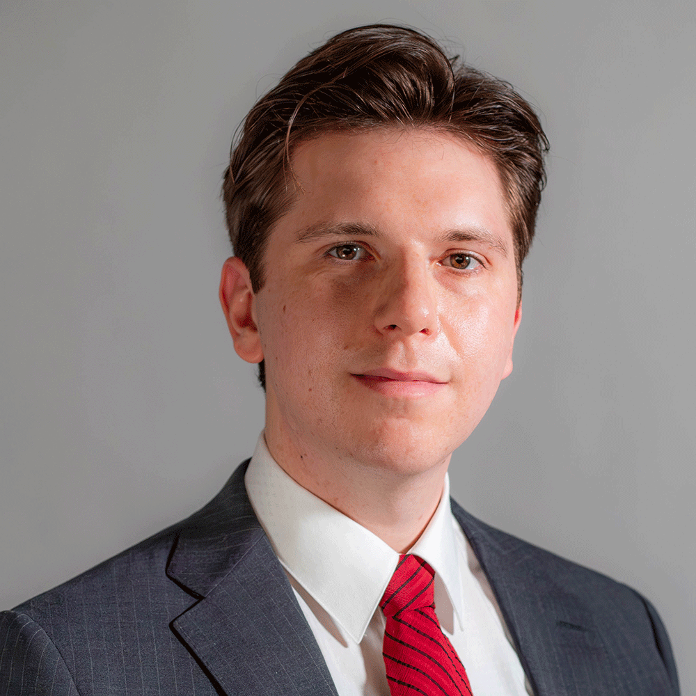 Headshot of a young man wearing a dark suit, white shirt, and red striped tie against a light gray background.