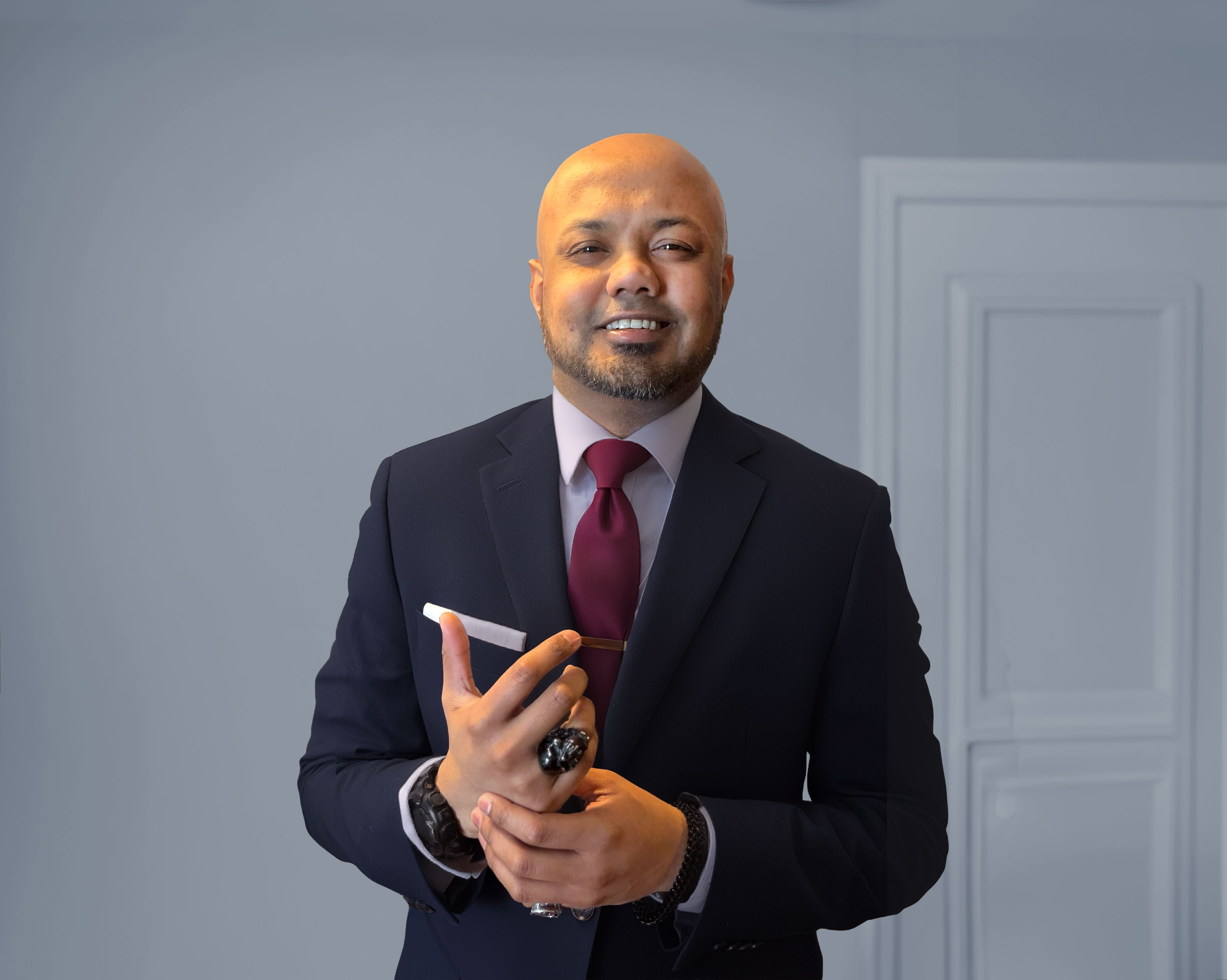 Headshot of a young man wearing a dark navy suit, light dress shirt, and burgundy tie, standing indoors with hands clasped.
