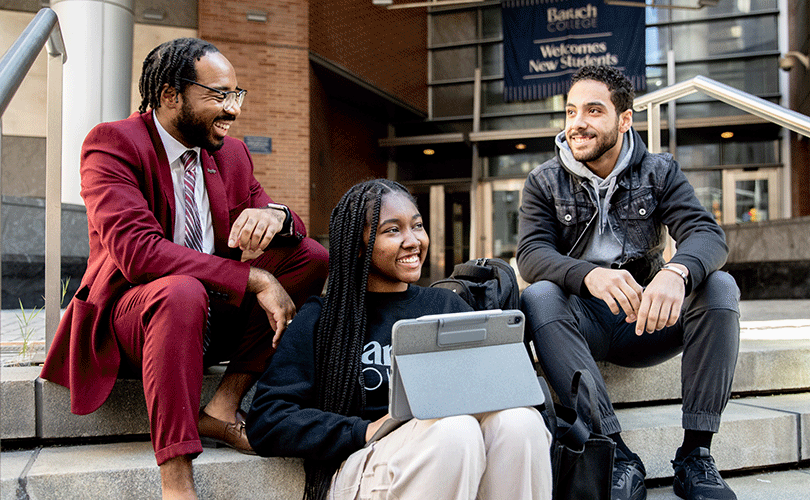 Three seated students talking on campus steps at Baruch College entrance. One holds a tablet. A&ldquo; Welcoming New Students&rdquo; banner hangs in the background.