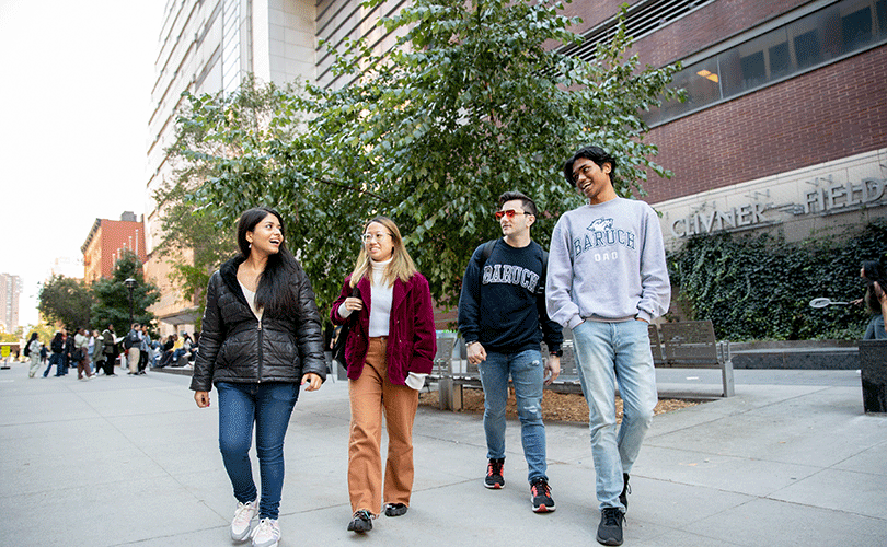 A group of four students walk through Baruch’s outdoor plaza on East 25th Street, passing trees and seating areas beside a large campus building.