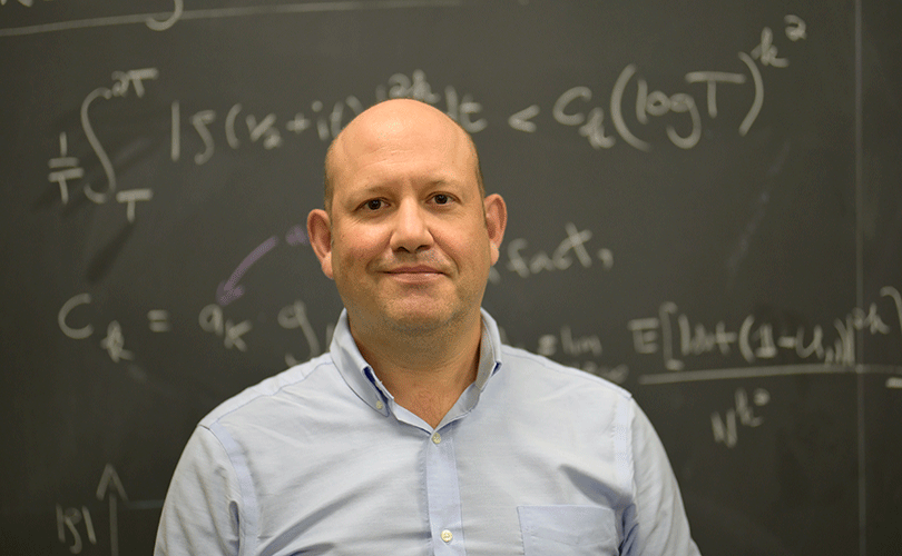 Headshot of Baruch Professor Louis-Pierre Arguin standing in front of a chalkboard covered with mathematical equations.